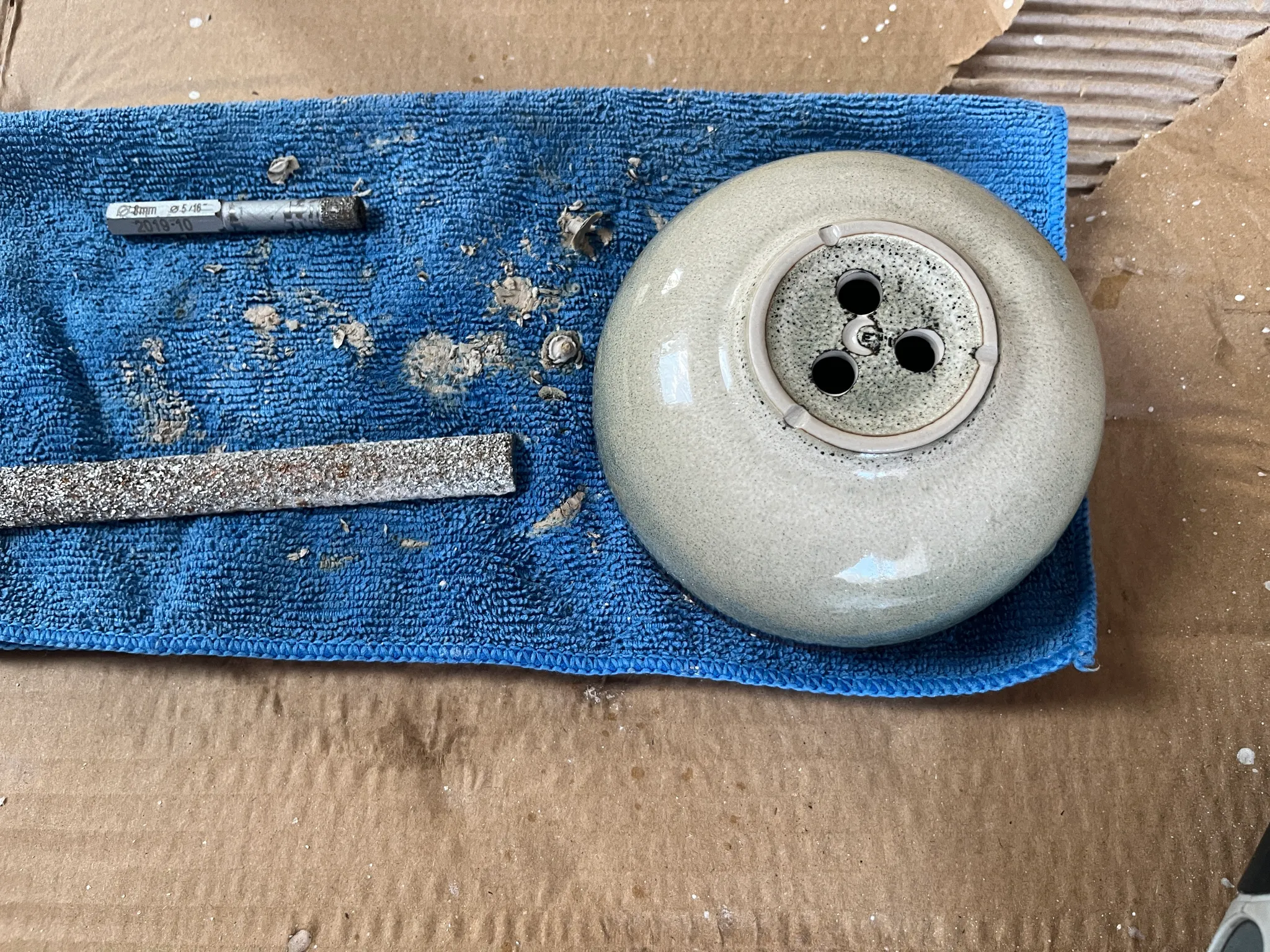 The underside of a grey glazed bonsai pot on a blue cloth over cardboard, showing three drainage holes, with a sharpening file beside it.