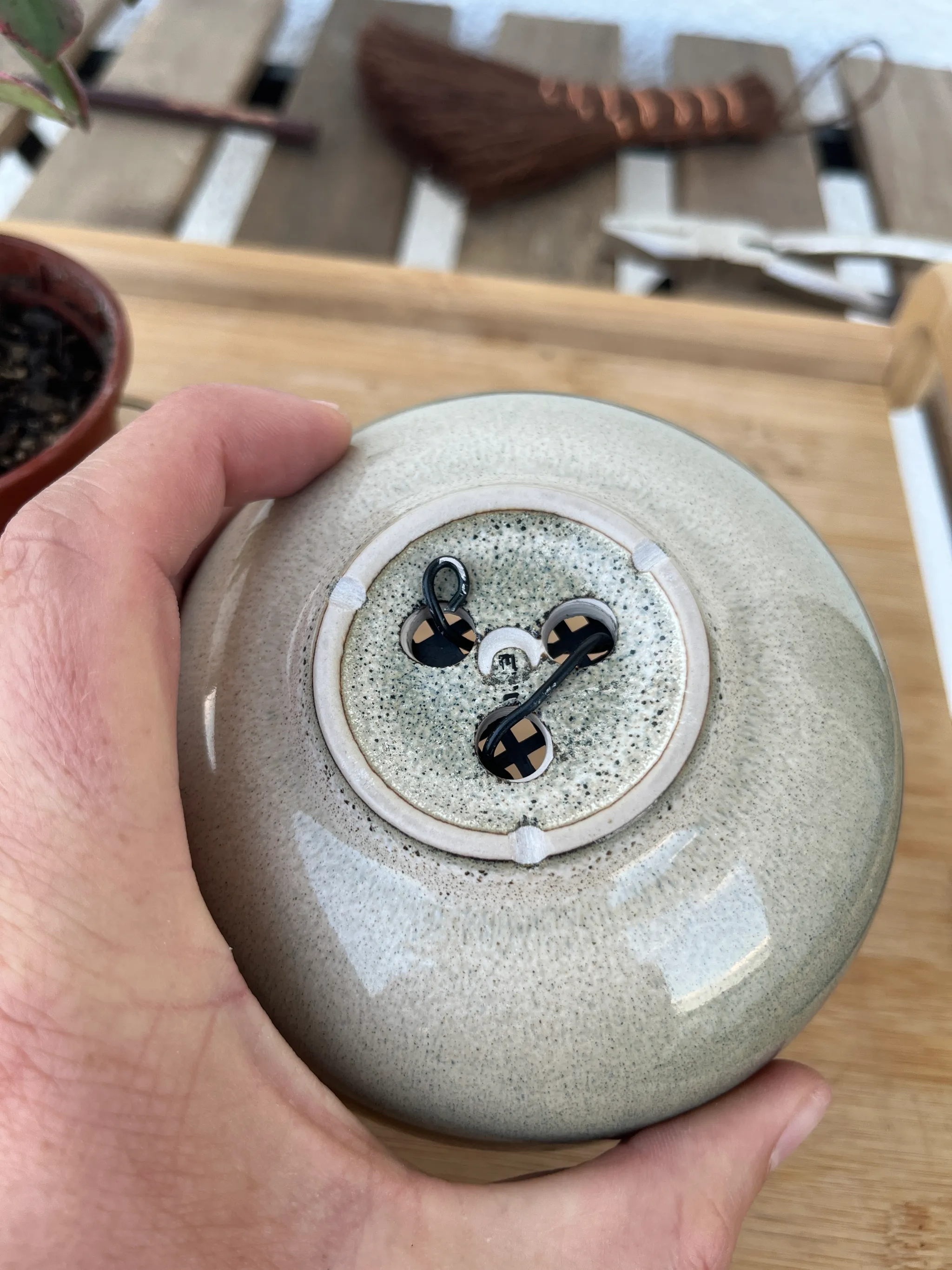 A hand holding the grey glazed bonsai pot from below, showing anchoring wire threaded through the drainage holes.